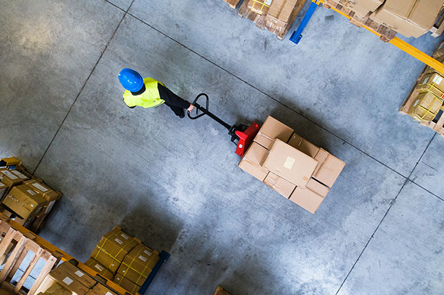 A human in a hi-vis vest and a blue hard hat works in a logistic centre moving a palette of boxes